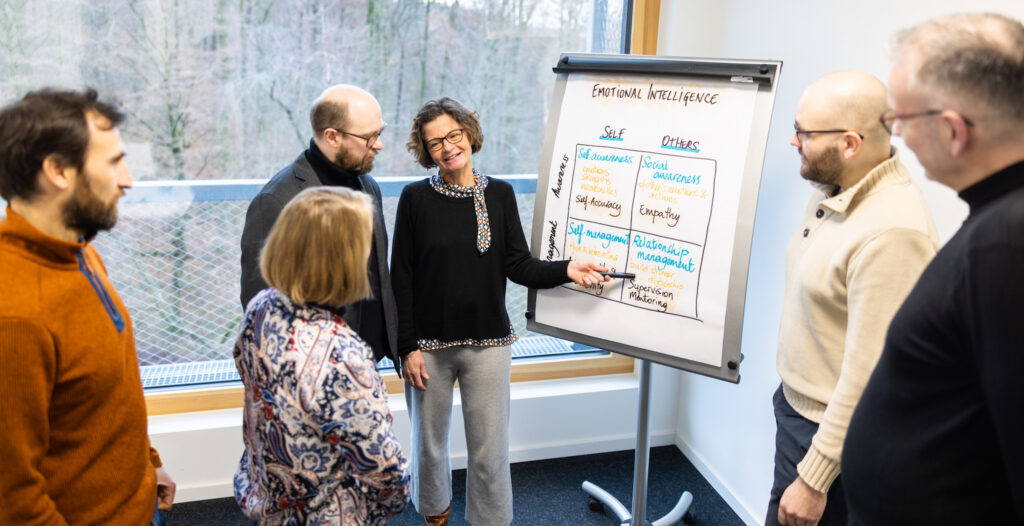 A group of people are standing around a flip chart showing a diagram of the four quadrants of Emotional Intelligence. A woman is standing at the flip chart presenting the information.
